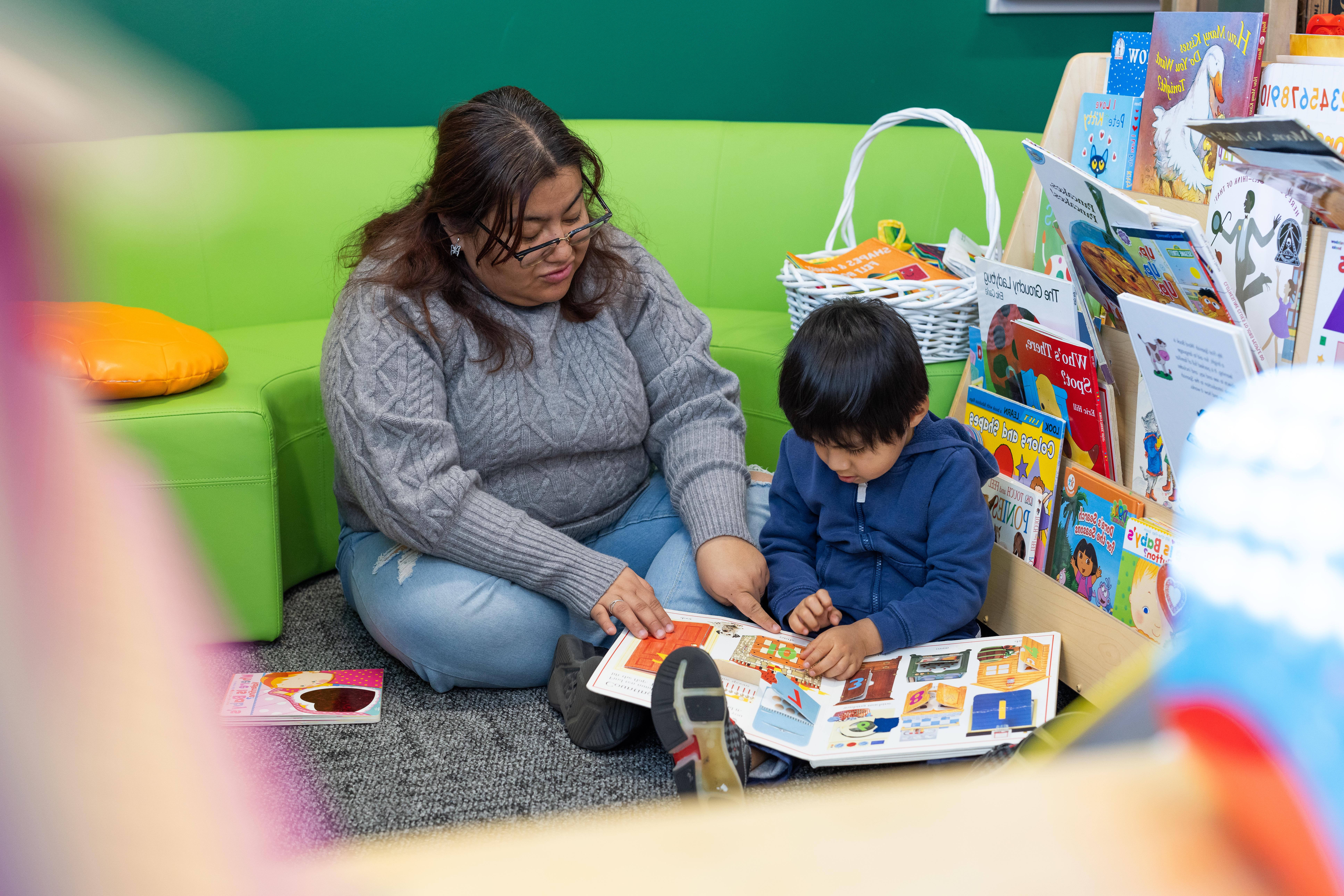 A woman sits on the floor with a young child reading a book next to a rack full of children's books.